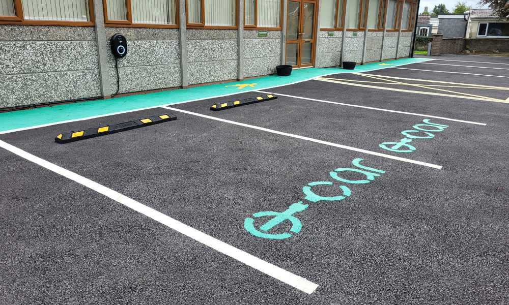 EV charging parking spots, with green writing and black and yellow wheel stops. Pedestrian walkway in the background.