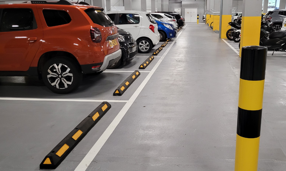 Underground car park with cars and motorcycles. Black and yellow wheel stops and bollards feature in the foreground.